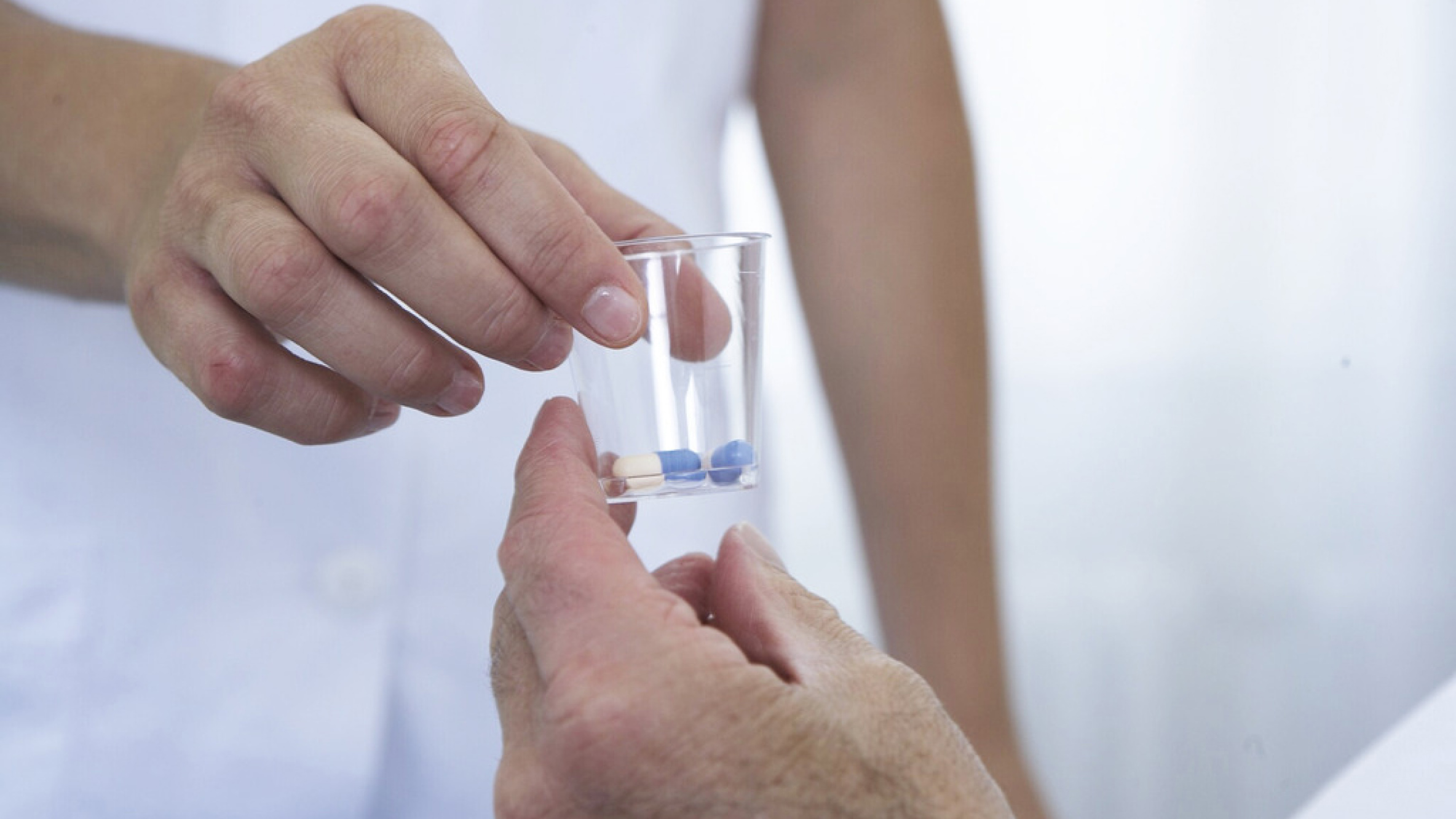 Eine Person gibt einer anderen Person ein kleines durchsichtiges Plastikglas mit mehreren Pillen darin.