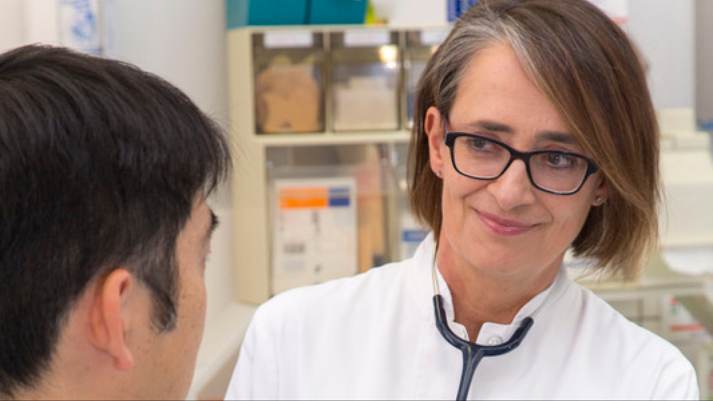 Médecin d'âge moyen regarde avec un léger sourire un homme qui reste dos au spectateur. En arrière-plan, on aperçoit plusieurs tiroirs contenant des éléments de la pratique médicale.